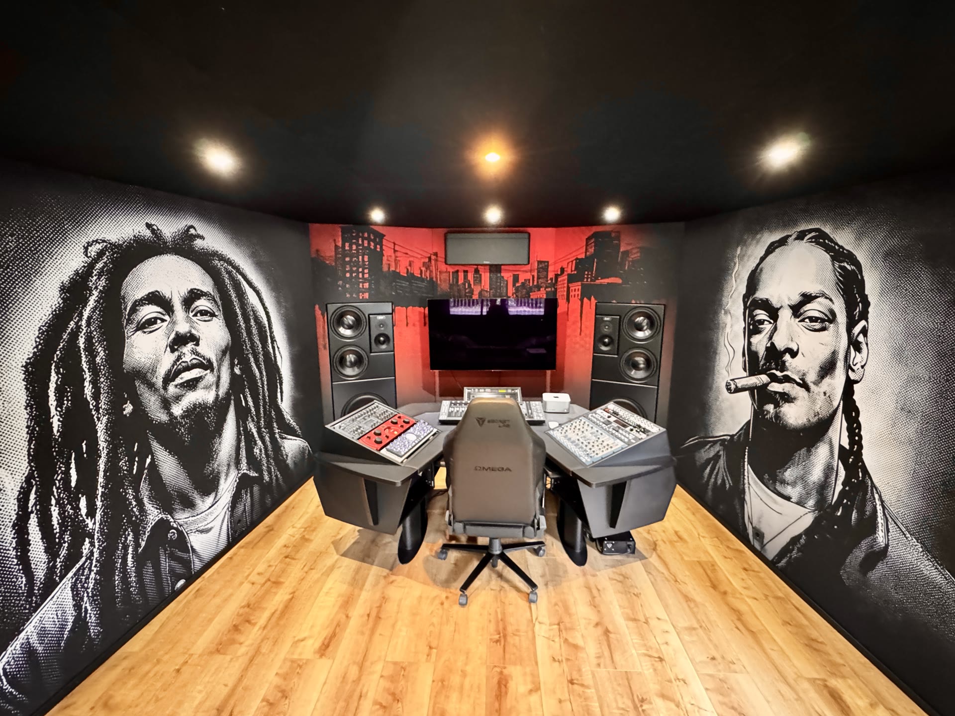 Daylight view of the MMA Studios control room: large-format graphic-novel-style portrait murals printed on acoustically transparent fabric covering the side walls, the red city-skyline artwork behind the monitor wall, warm wood floor.