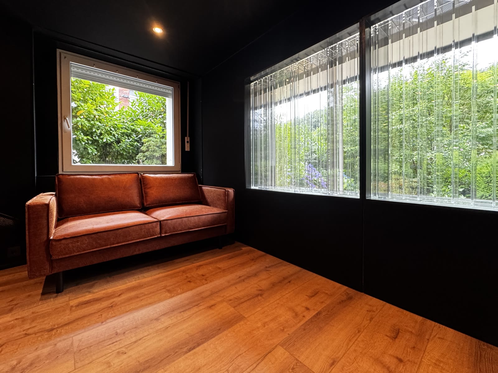 The lounge corner: cognac leather sofa on warm wood flooring, daylight falling through the Lightffusor windows with the garden behind.
